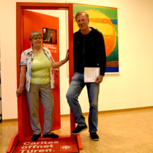 Rosemarie Horlemann und Stefan Weber stehen mit dem unterschriebenen Geschäftsbesorgungsvertrag in der roten Caritastüre.